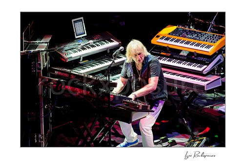 Horizontal color image of Geoff Downes of Yes performing at a large keyboard rig surrounded by multiple synthesizers during a live performance