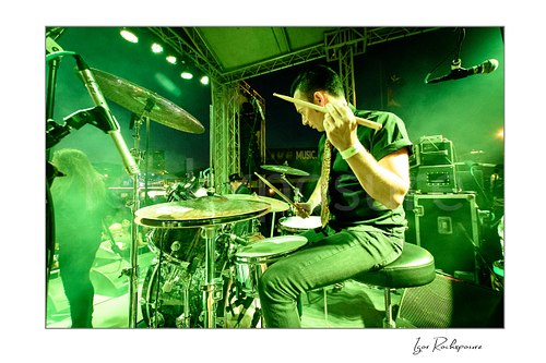 Horizontal color image of Laust Sonne playing drums on stage under vivid green concert lighting, with drumsticks raised and stage equipment visible