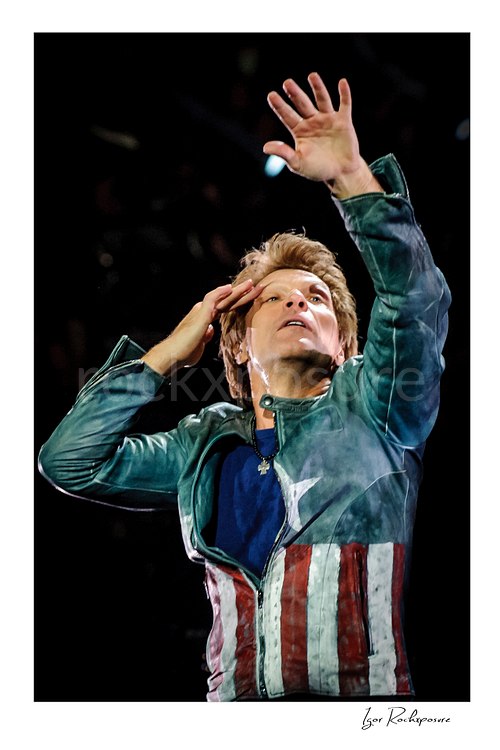 Vertical color image of Jon Bon Jovi in a dramatic live performance close-up, raising one hand while the other rests near his face against a dark stage background