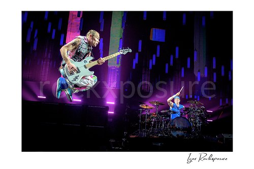 Flea leaps high with his white bass and knees tucked while Chad Smith raises a drumstick at his kit beneath hanging LEDs and purple light.