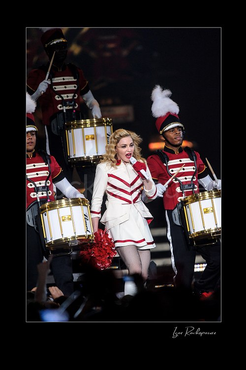 Vertical color image of Madonna singing into a microphone in a white and red stage outfit while marching drummers perform around her