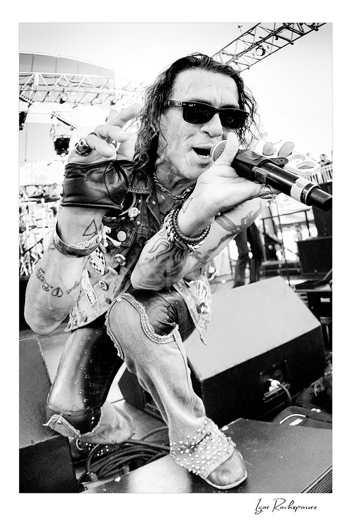 Vertical black and white image of Stephen Pearcy of Ratt singing into a microphone in a tight stage-level close-up while wearing sunglasses