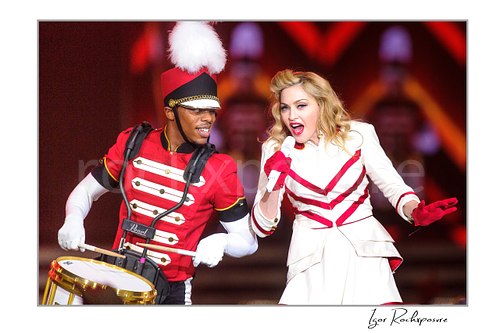 Horizontal color image of Madonna performing beside a drummer in marching band costume during a theatrical live stage scene