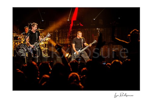 Horizontal color image of Chad Kroeger and Mike Kroeger of Nickelback performing onstage with the drummer visible behind them and a silhouetted crowd in front