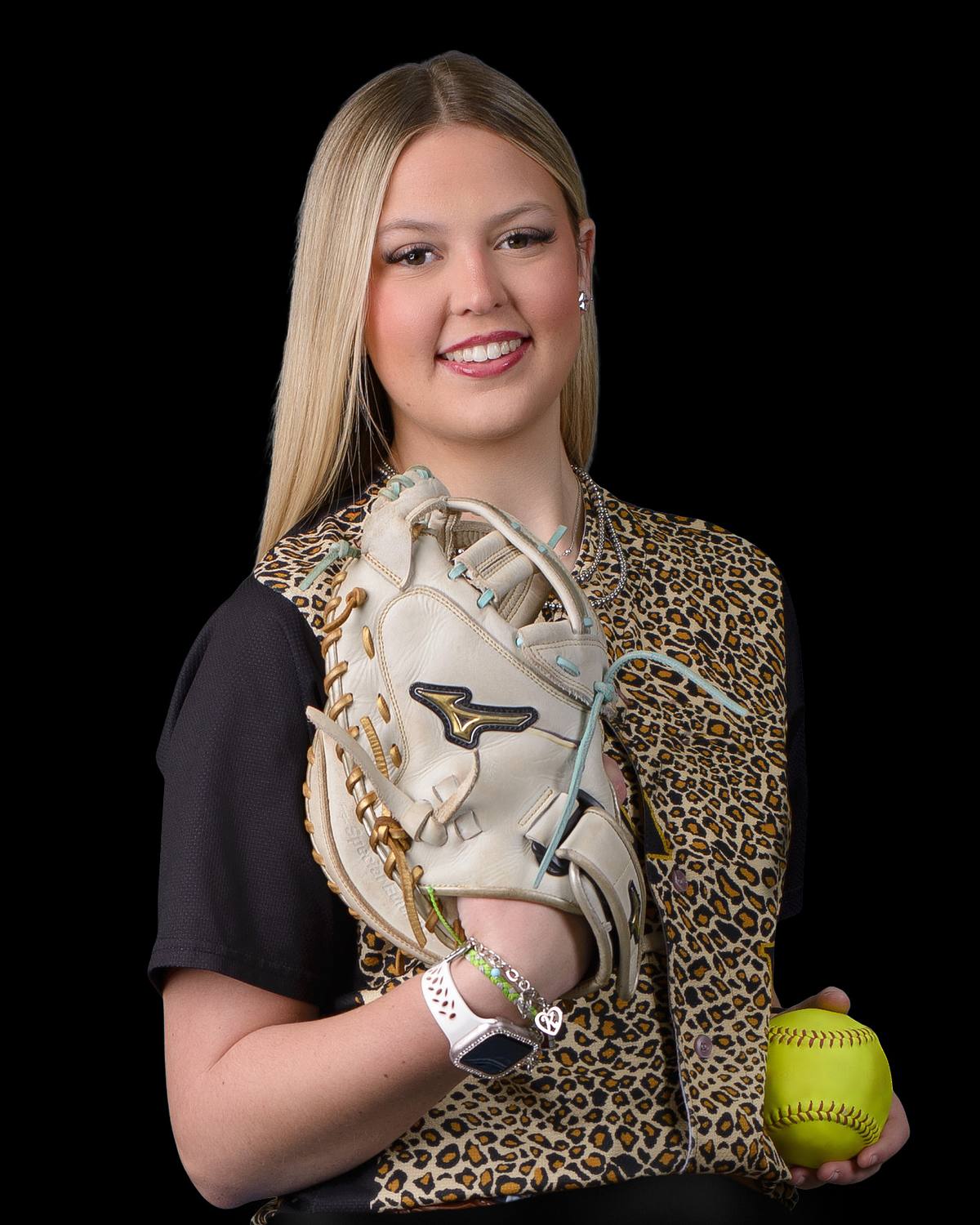 A smiling young female high school softball player with long blonde hair, wearing a leopard print shirt. She holds a cream-colored Mizuno softball glove in her left arm and a bright yellow softball in her right hand, against a pure black background. | ProClicks Photography