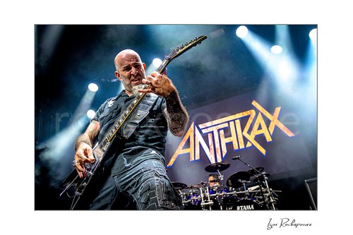 Horizontal color image of Scott Ian of Anthrax performing live on stage, playing guitar in front of the Anthrax stage backdrop under bright concert lighting