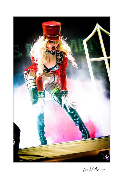 Vertical color image of Maria Brink of In This Moment in a theatrical live performance pose, wearing a red military-style outfit with tall black boots amid stage smoke and bright backlight