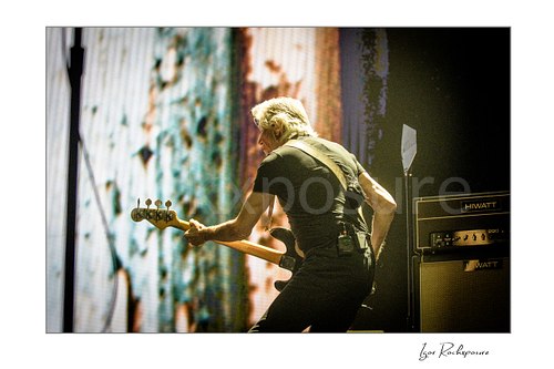 Roger Waters plays bass with his back turned toward the camera, its wooden neck extending left beside an amplifier and a brightly patterned stage screen.
