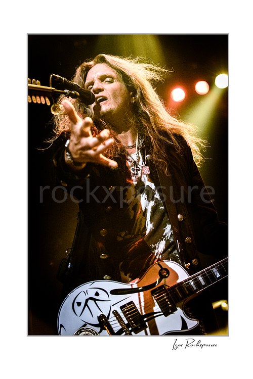 Vertical color close-up image of Joel Hoekstra of Whitesnake performing live on stage, singing into a microphone while playing electric guitar under warm concert lighting