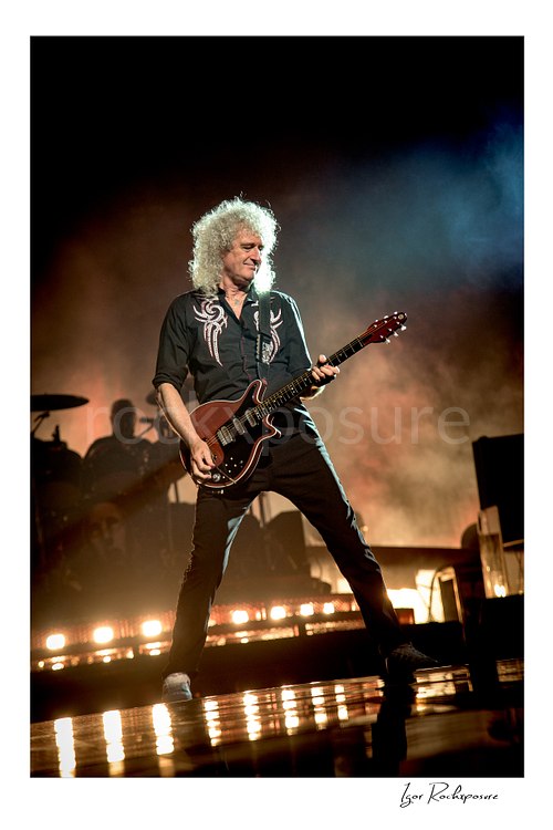 Vertical color image of Brian May of Queen playing guitar onstage in a wide stance with glowing stage haze and warm floor lights
