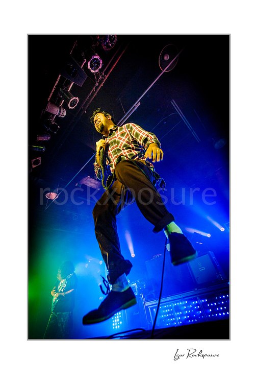 Vertical color image of Chino Moreno of Deftones performing live on stage in a plaid shirt with a handheld microphone, airborne jump, and vivid blue-green lighting