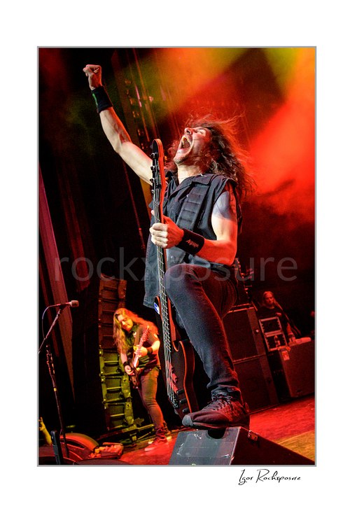 Vertical color image of Frank Bello of Anthrax performing live on stage, raising one fist while holding his bass guitar under vivid red and yellow concert lighting