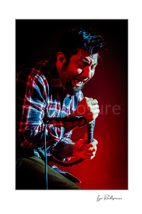 Vertical color image of Chino Moreno of Deftones performing live on stage with a handheld microphone, plaid shirt, intense expression, and deep red lighting