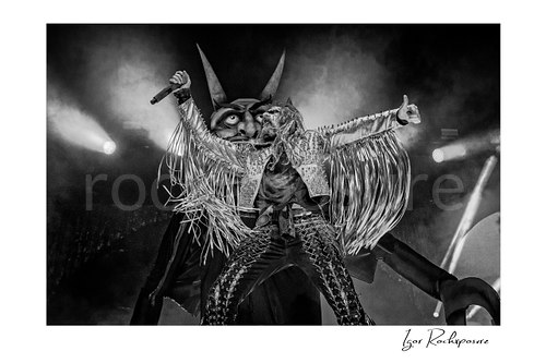 Horizontal black and white image of Rob Zombie performing live on stage in a fringed jacket with raised arms, handheld microphone, and a giant horned stage figure behind him