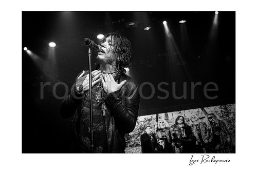 Horizontal black and white image of Tom Keifer singing at the microphone in an emotional live performance moment, with both hands at his chest under dramatic stage lighting and a band image visible behind him