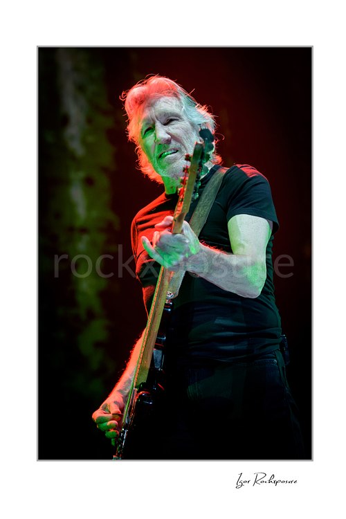 Roger Waters angles his bass neck toward the camera while playing, with green light across his face and arms and red light in his hair.