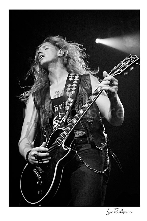 Vertical black and white image of Jared James Nichols playing guitar onstage under a bright spotlight