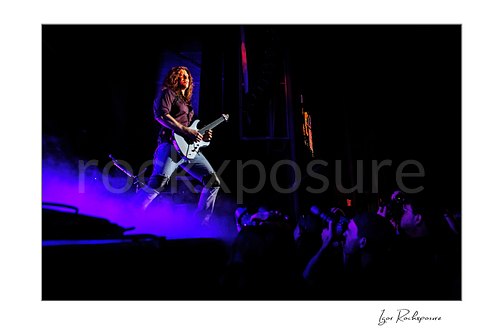 Horizontal color image of Kiko Loureiro of Megadeth playing guitar on stage above purple smoke with fans visible below