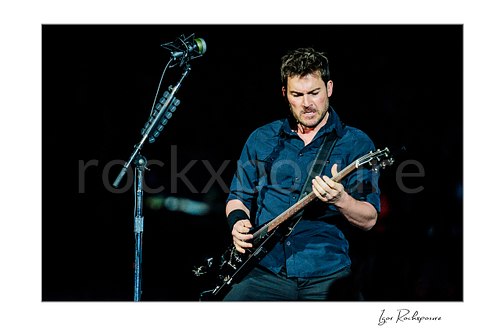 Horizontal color image of Ryan Peake of Nickelback playing guitar onstage beside a microphone stand in a tight live performance shot