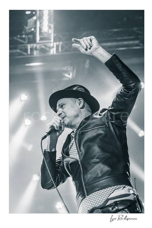 Vertical black and white image of Gord Downie of The Tragically Hip singing into a microphone with one arm raised, wearing a hat and leather jacket under bright stage lights
