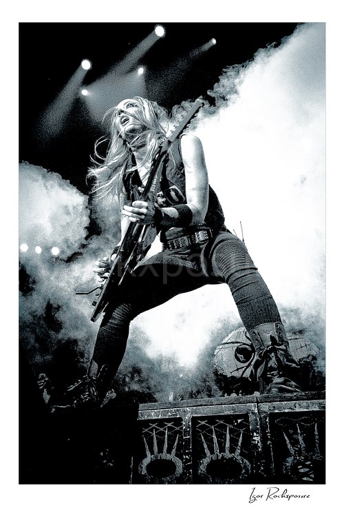 Vertical black and white image of Nita Strauss performing live on stage with an electric guitar, surrounded by thick stage smoke