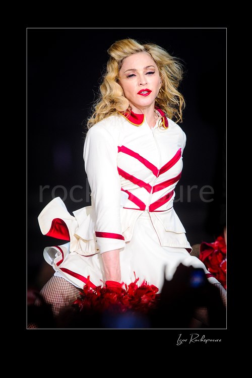 Vertical color image of Madonna in a white and red stage outfit during a theatrical live performance with red pom-poms visible in the foreground