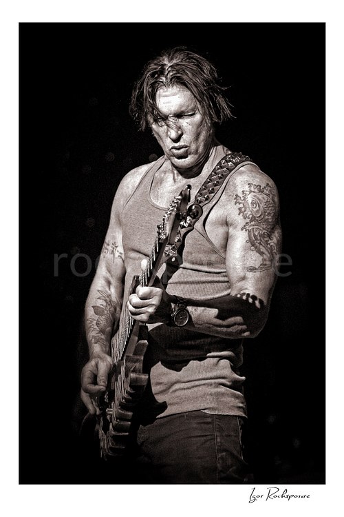 Vertical black and white image of George Lynch playing guitar on stage with a focused expression and tattooed arm