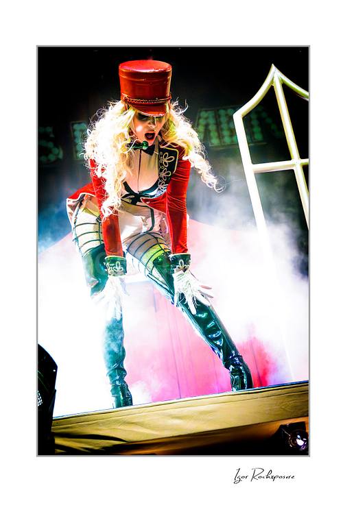 Vertical color image of Maria Brink of In This Moment in a theatrical live performance pose, wearing a red military-style outfit with tall black boots amid stage smoke and bright backlight