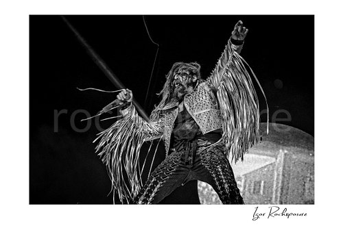 Horizontal black and white image of Rob Zombie performing live on stage in a fringed jacket with raised arm, dreadlocks, and a handheld microphone