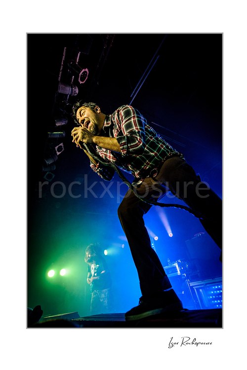 Vertical color image of Chino Moreno of Deftones performing live on stage in a plaid shirt with a handheld microphone, low-angle perspective, and blue-green concert lighting