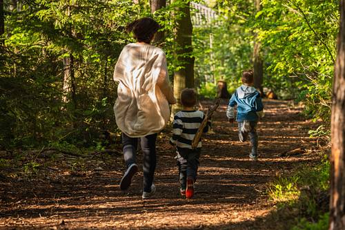 Eine Mutter und ihre zwei kleinen Söhne rennen voller Energie von hinten gesehen einen sonnendurchfluteten Waldweg entlang.