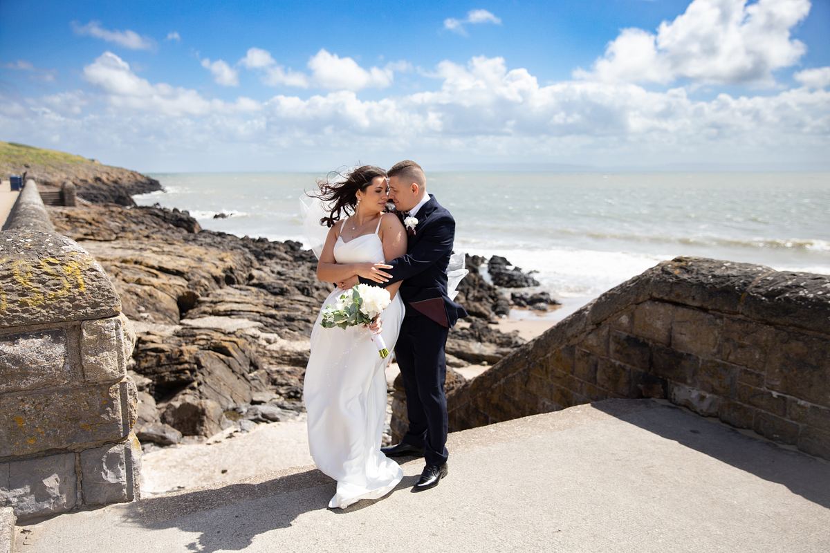 Bride and groom embracing beside the rocky coast at Barry Island, South Wales
