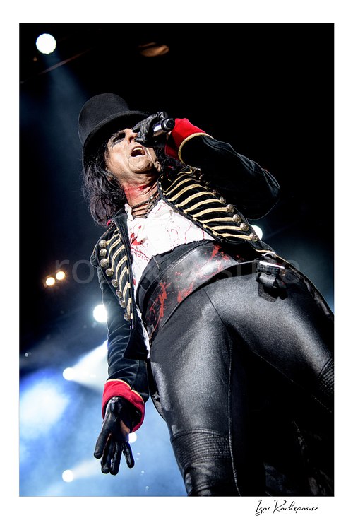 Vertical color image of Alice Cooper performing live on stage in a black top hat, military-style jacket, blood-stained shirt, and black leather pants under bright stage lights