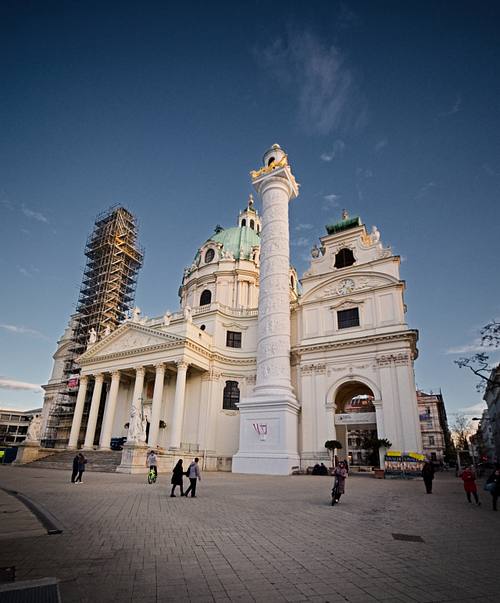 Barocke Kirche mit großer Kuppel und Siegessäule auf einem weitläufigen Platz in Wien bei blauem Abendhimmel.