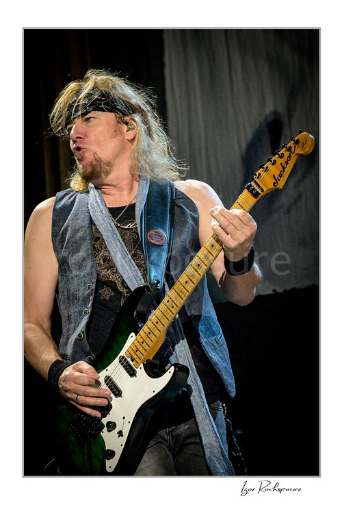 Vertical color image of Adrian Smith of Iron Maiden performing live on stage, playing electric guitar while wearing a bandana and sleeveless denim vest