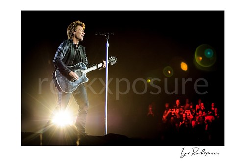 Horizontal color image of Jon Bon Jovi performing live with an acoustic guitar beside a microphone stand, backlit by a bright spotlight with lens flare and audience visible in the distance