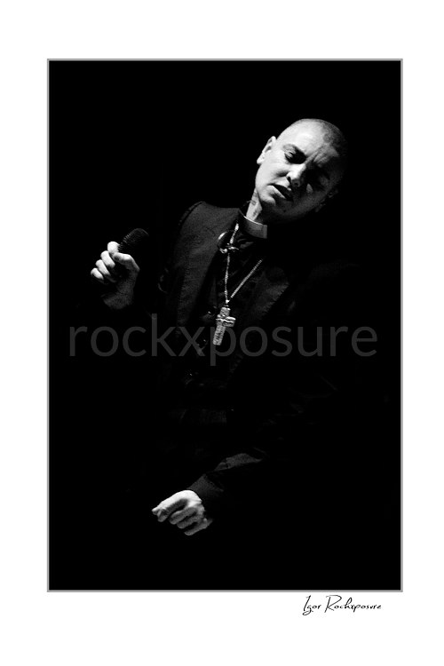 Sinéad O’Connor tilts her head while holding a microphone near her shoulder, wearing a dark jacket and cross necklace in a black and white portrait.
