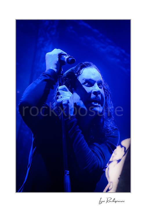 Ozzy Osbourne of Black Sabbath holding a microphone above his head during a live performance under deep blue concert lighting