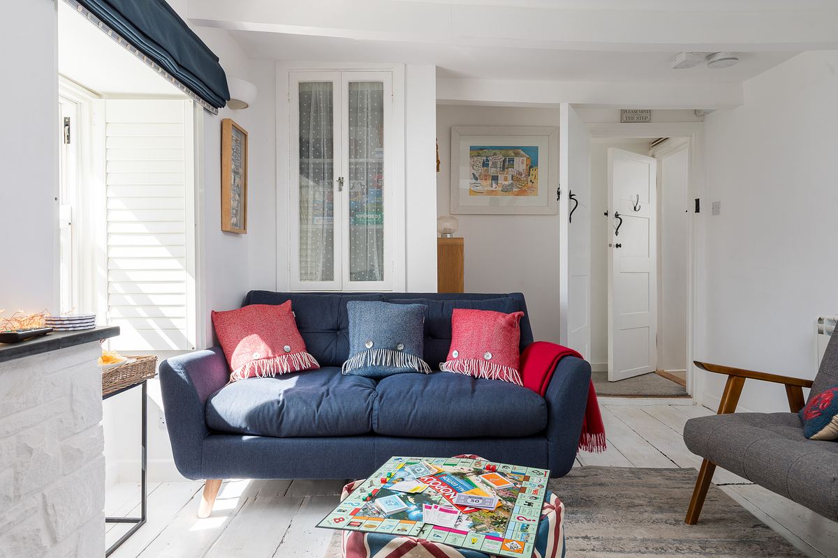 A small, bright living room in St Ives features a blue sofa with colorful pillows, a patterned coffee table, and light-colored walls with artwork and a few doors visible in the background.