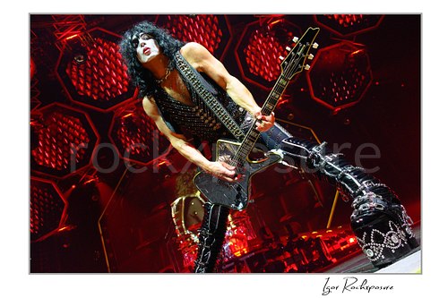 Paul Stanley of KISS performing a wide power stance while playing guitar under intense red stage lighting with dramatic circular stage visuals