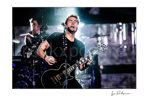 Horizontal color image of Chad Kroeger of Nickelback playing guitar onstage beside a microphone stand with the drummer visible behind him under bright backlight