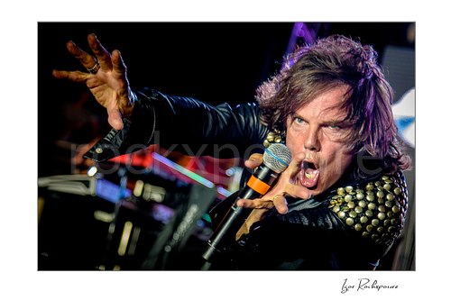 Horizontal color image of Joey Tempest of Europe performing live on stage with a handheld microphone, black leather jacket with studded shoulders, intense expression, and outstretched hand