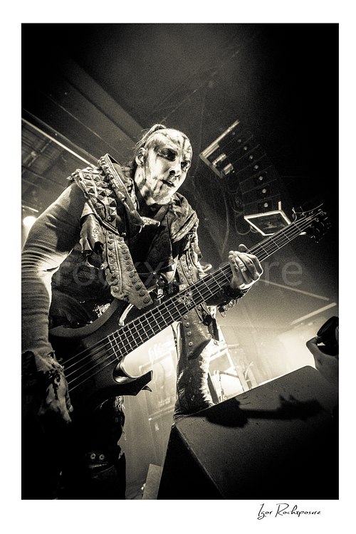 Vertical black and white image of Orion of Behemoth playing bass in a dramatic low-angle live performance shot with corpse paint and stagewear