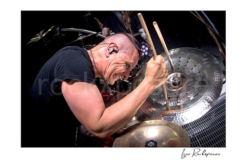 Horizontal color image of Korn drummer Ray Luzier in a dramatic close portrait with a drumstick pressed against a cymbal, a scary expression, and bright stage lighting