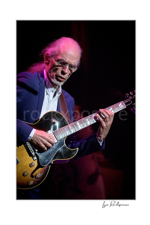 Vertical color image of Steve Howe of Yes playing guitar onstage under red and purple concert light