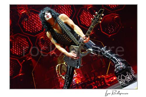 Paul Stanley of KISS performing a wide power stance while playing guitar under intense red stage lighting with dramatic circular stage visuals