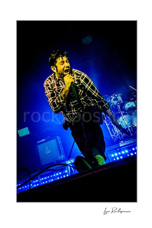 Vertical color image of Chino Moreno of Deftones performing live on stage with a handheld microphone, plaid shirt, blue stage lighting, and the drum kit visible behind him