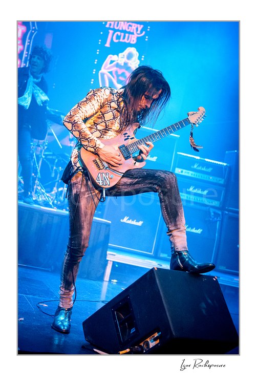 Vertical color image of Nuno Bettencourt performing live on stage with an electric guitar, one foot raised on a stage monitor under vivid blue lighting