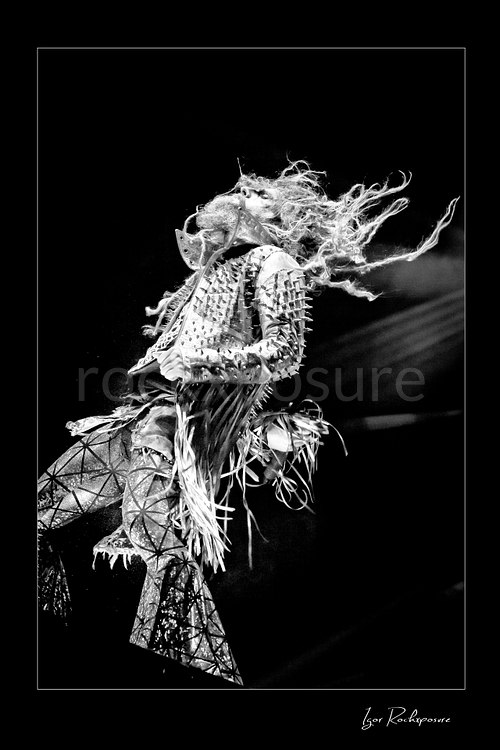 Vertical black and white image of Rob Zombie performing live on stage with flying dreadlocks, a studded fringed jacket, and dramatic backward motion