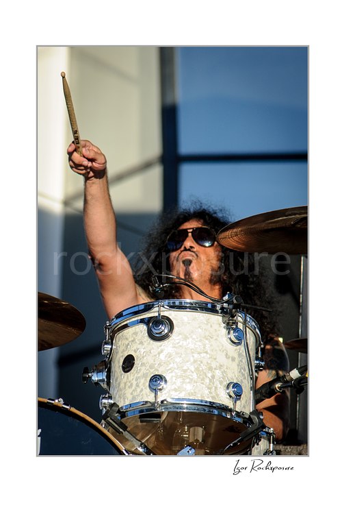 Vertical color image of Frankie Banali behind a white drum kit with one drumstick raised high during a live performance